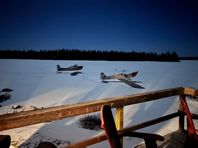 A night photo, brightly lit, of two planes on skis on the ice.