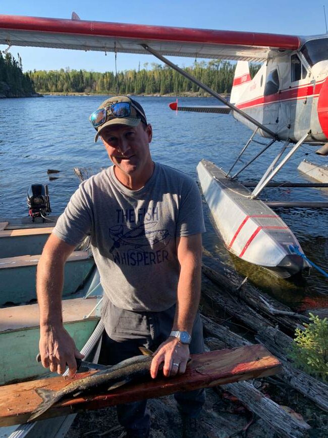 Chris fileting fresh caught lake trout on a board with the boat and float plane in the background.