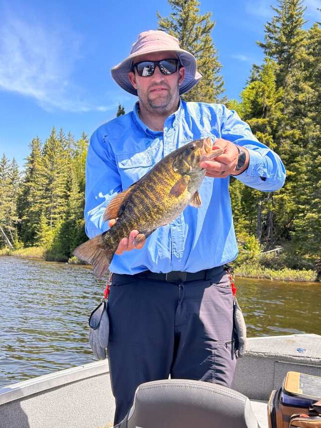 Fisherman holding a smallmouth bass fishing on a beautiful day with green trees in the background.